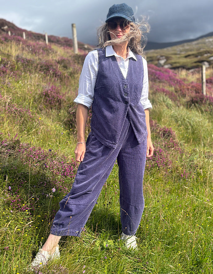 A woman stands outdoors in a grassy field with purple flowers and hills in the background, wearing a matching blue corduroy vest and trousers set over a white collared shirt, with a blue cap and white shoes.