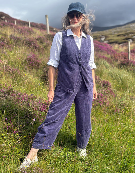 A woman stands outdoors in a grassy field with purple flowers and hills in the background, wearing a matching blue corduroy vest and trousers set over a white collared shirt, with a blue cap and white shoes.