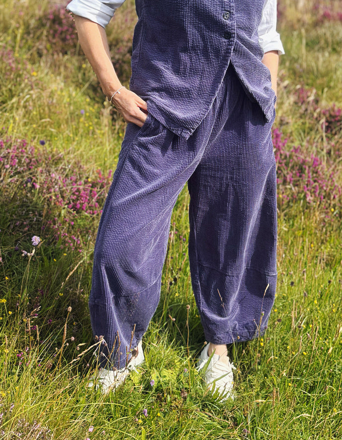 A person standing in a grassy field with wildflowers, wearing loose dark blue corduroy pants and a matching vest over a light blue shirt, with white sneakers.