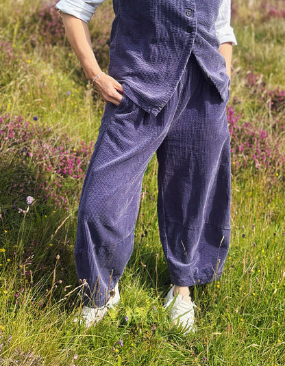 A person standing in a grassy field with wildflowers, wearing loose dark blue corduroy pants and a matching vest over a light blue shirt, with white sneakers.