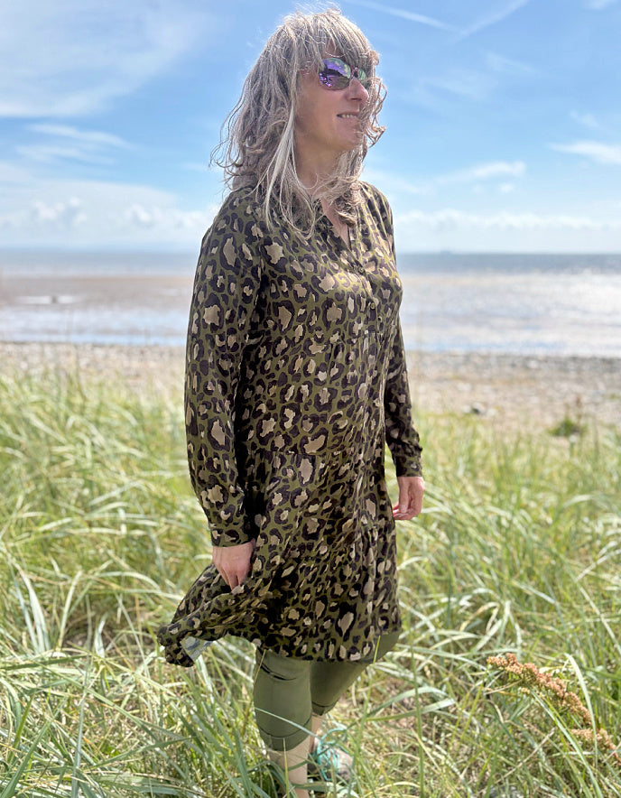 Person wearing a leopard print dress standing on a grassy field with a beach and blue sky in the background