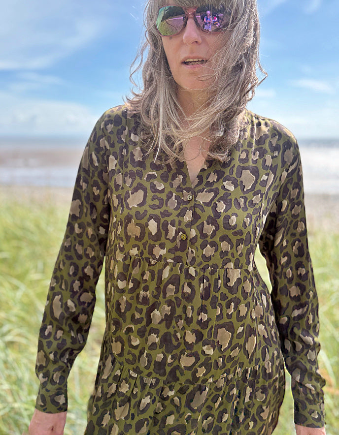 Person wearing a leopard print dress standing on a grassy field with a beach and blue sky in the background