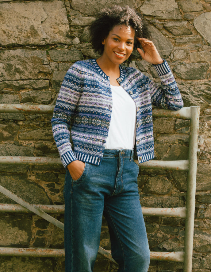 Woman wearing a blue patterned cardigan and jeans standing against a stone wall.