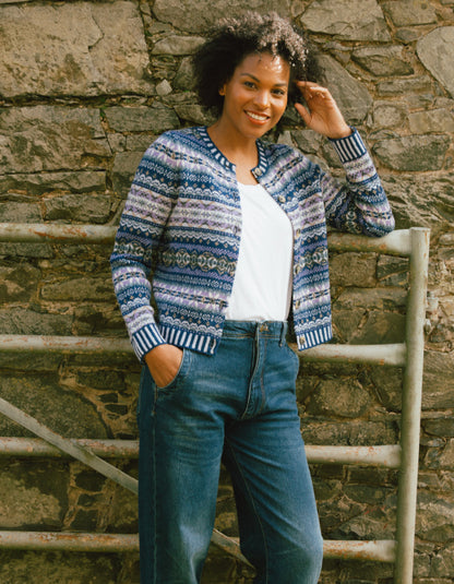 Woman wearing a blue patterned cardigan and jeans standing against a stone wall.