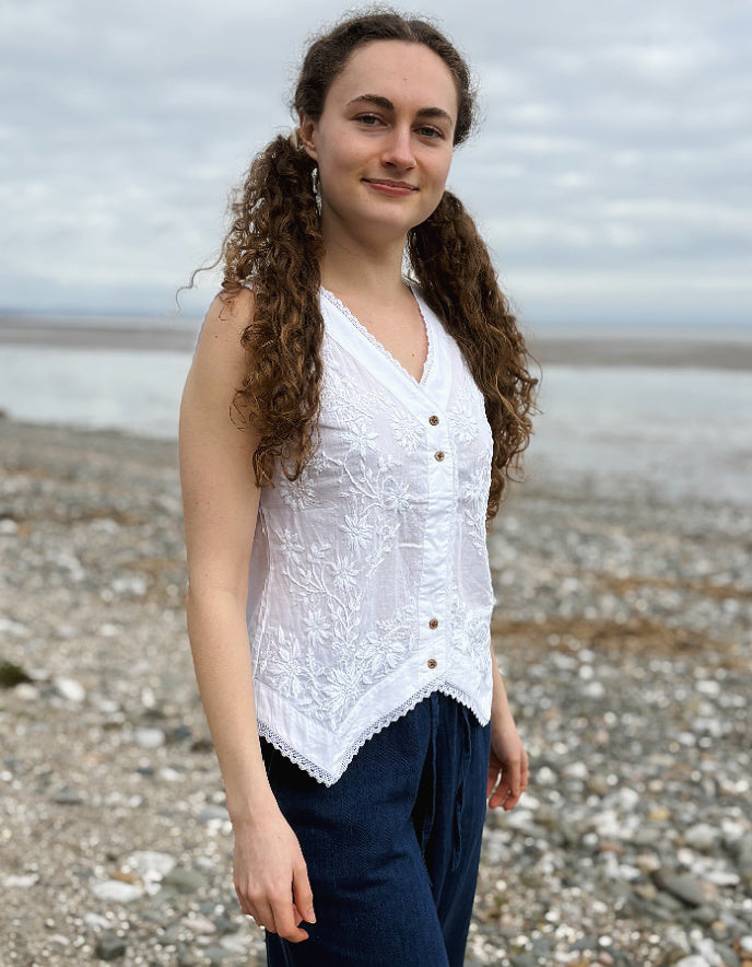 Woman wearing a white lace top and navy pants standing on a pebbly beach.