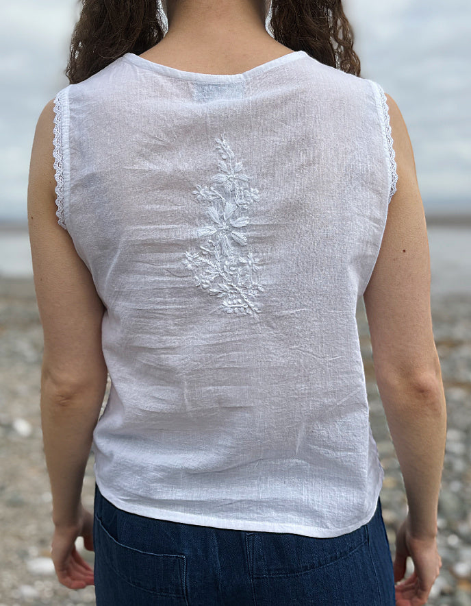 Person wearing a white sleeveless top with lace trim at the neckline, standing on a beach.