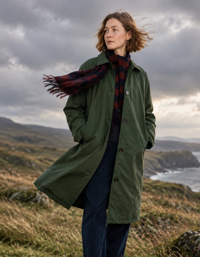 A woman stands outdoors in a grassy coastal landscape wearing an olive green trench coat over a dark red and navy plaid scarf and dark blue trousers, with a cloudy sky overhead.