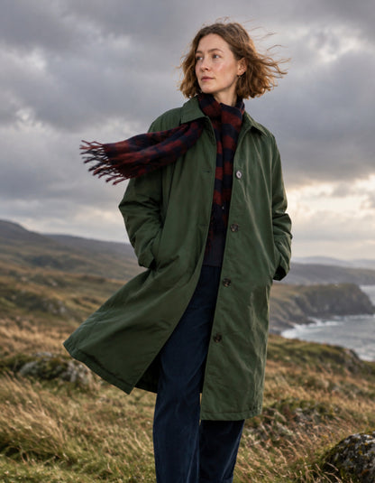 A woman stands outdoors in a grassy coastal landscape wearing an olive green trench coat over a dark red and navy plaid scarf and dark blue trousers, with a cloudy sky overhead.