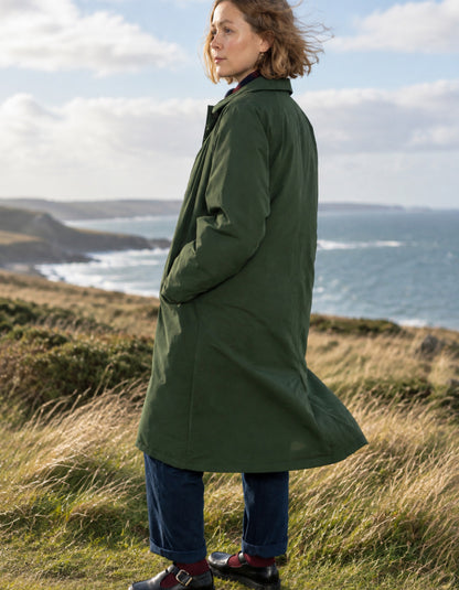 A woman wearing a long dark green coat, blue jeans, and brown shoes stands on a grassy coastal hillside with the ocean and cloudy sky in the background.