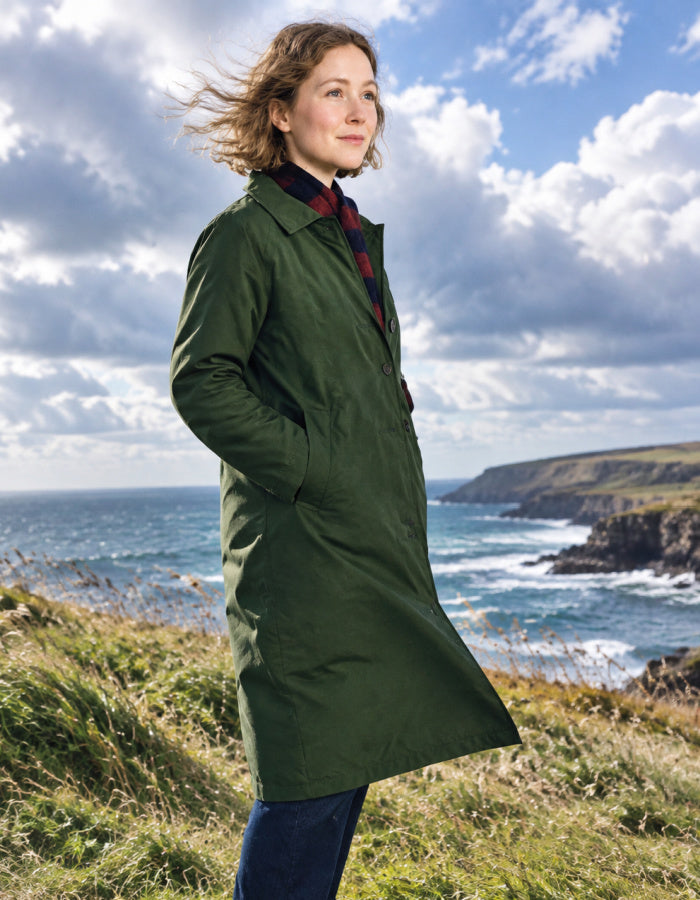 A woman stands on a grassy coastal cliff wearing a long olive green coat over a red plaid shirt and dark pants, with the ocean and cloudy sky behind her.