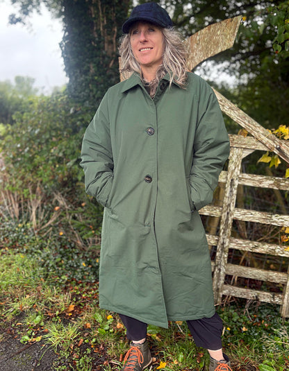 A woman wearing an olive green mid-length coat, black pants, and a dark cap stands outdoors in front of a rustic wooden fence and greenery.