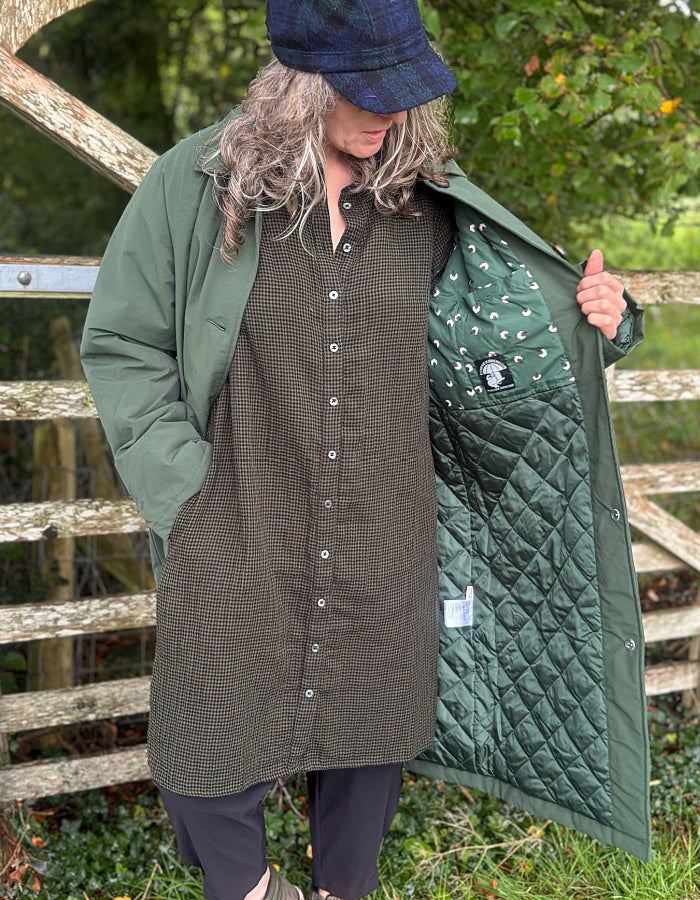 A person outdoors wearing an olive green quilted coat over a matching green button-front dress, with the coat open to show a green quilted lining. A wooden fence and greenery are in the background.