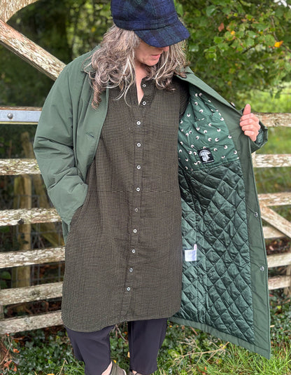 A person outdoors wearing an olive green quilted coat over a matching green button-front dress, with the coat open to show a green quilted lining. A wooden fence and greenery are in the background.
