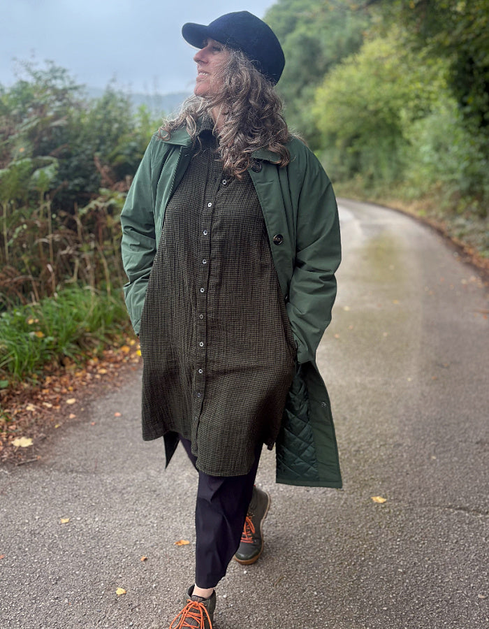 A woman walking on a paved country path wearing a dark green quilted coat over a dark olive button-front dress, with black pants and orange sneakers. She is also wearing a black beret.