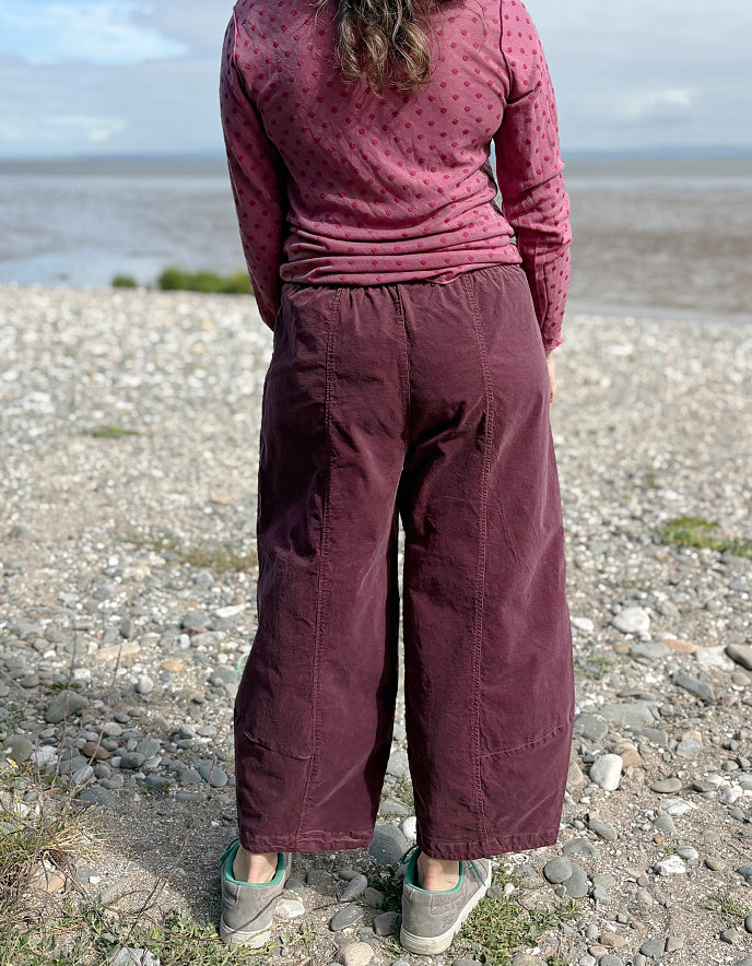 Person wearing a pink long-sleeve top and purple pants standing on a pebbly beach.