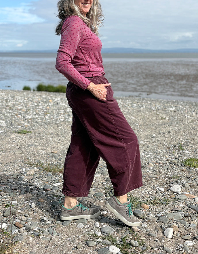 Woman in pink polka dot top and maroon pants standing on a rocky beach with water and sky in the background
