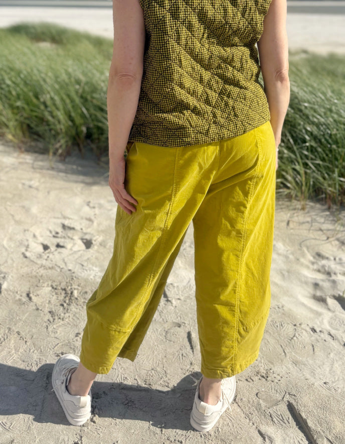 A person standing on a sandy beach wearing loose mustard yellow cropped pants, a textured olive green sleeveless top, and white sneakers, with dune grass in the background.