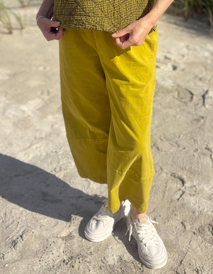 A person outdoors wearing loose mustard yellow cropped pants, a black-and-white checkered top, and white sneakers on sandy ground.