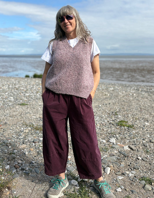 Person standing on a pebbly beach wearing a sleeveless pink sweater and maroon lantern pants.