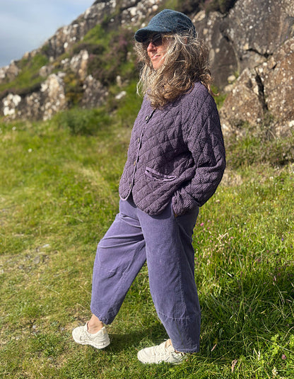 A woman stands outdoors on a grassy hillside wearing a quilted lavender cardigan, matching wide-leg trousers, a dark blue cap, and white sneakers.