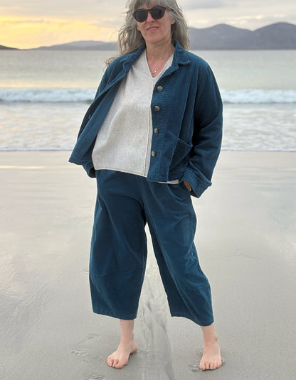 A woman stands barefoot on a beach wearing a dark teal blue corduroy jacket and matching cropped pants over a cream knit sweater, with ocean waves and mountains in the background.