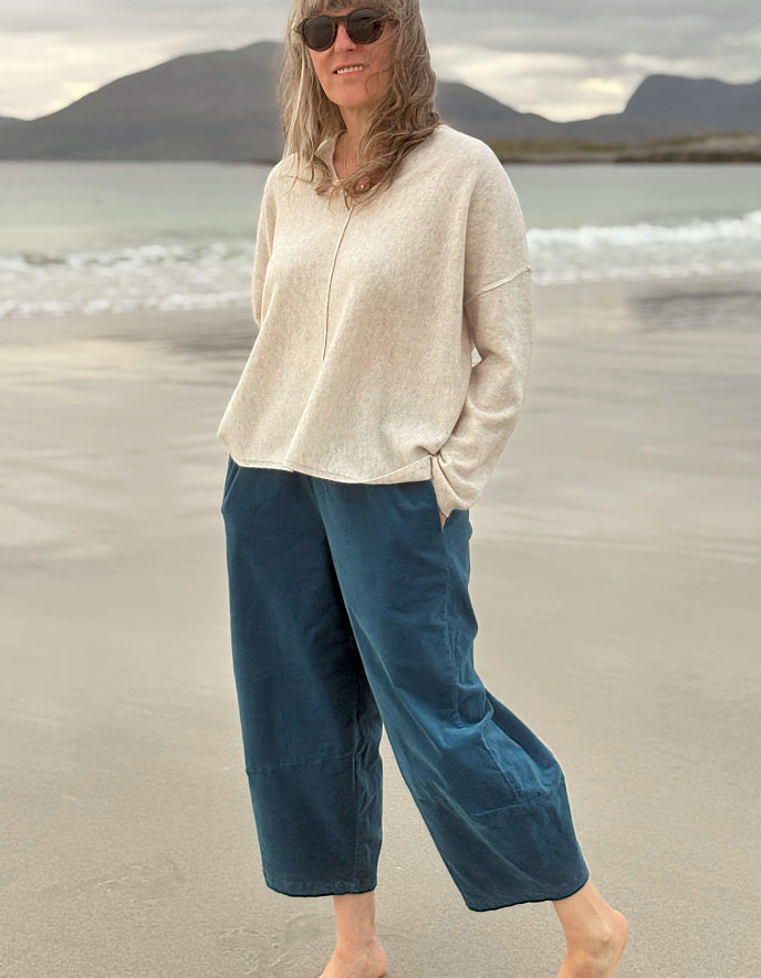 A woman standing on a sandy beach wearing a cream-colored knit top and dark teal wide-leg pants, with ocean waves and mountains in the background.