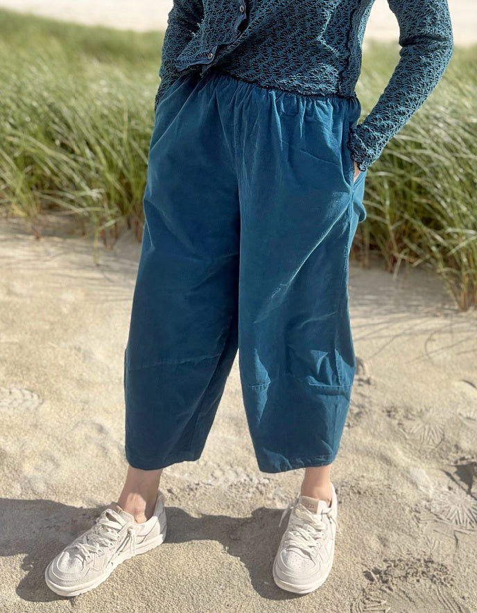 A person standing on sandy ground near beach grass, wearing loose teal-blue cropped pants, a patterned dark blue long-sleeve top, and white sneakers.