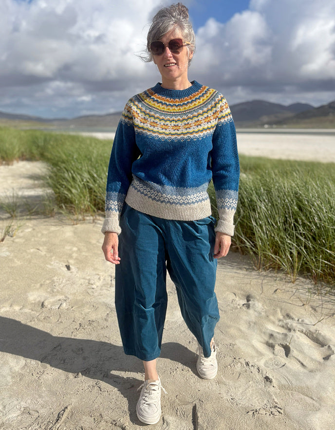 A woman standing outdoors on a sandy path near grassy dunes, wearing a blue knit sweater with a multicolor Fair Isle yoke and beige cuffs and hem, paired with teal cropped pants and white sneakers.