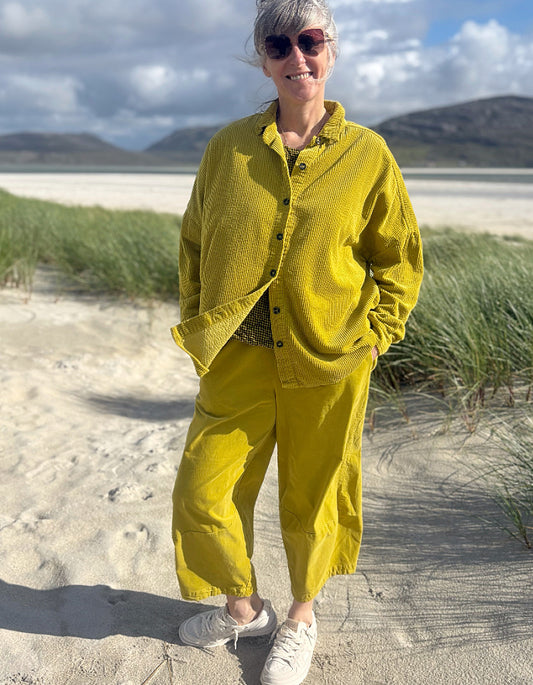 A woman standing on a sandy beach wearing a matching mustard yellow corduroy button-up shirt and trousers set, with grass, water, and mountains in the background.