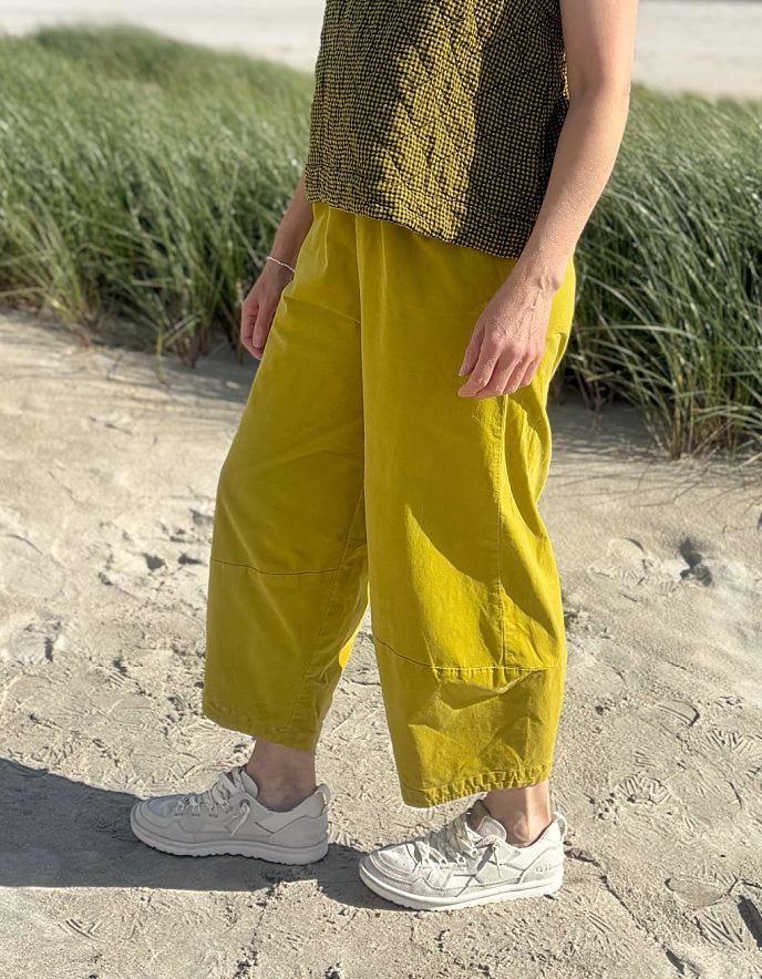 A person standing on a sandy beach wearing loose, wide-leg mustard yellow pants, a textured olive top, and white sneakers, with beach grass in the background.