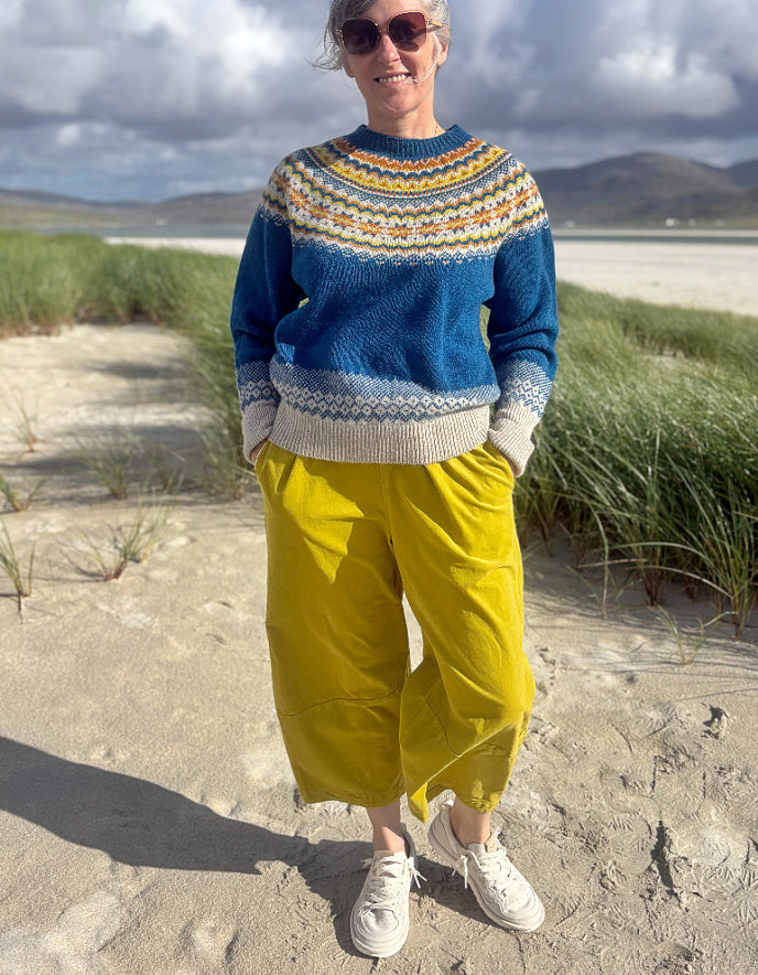 A woman standing outdoors on a sandy path near grassy dunes and water, wearing a blue Fair Isle knit sweater with cream, yellow, and orange patterning, bright yellow pants, white sneakers, and sunglasses.