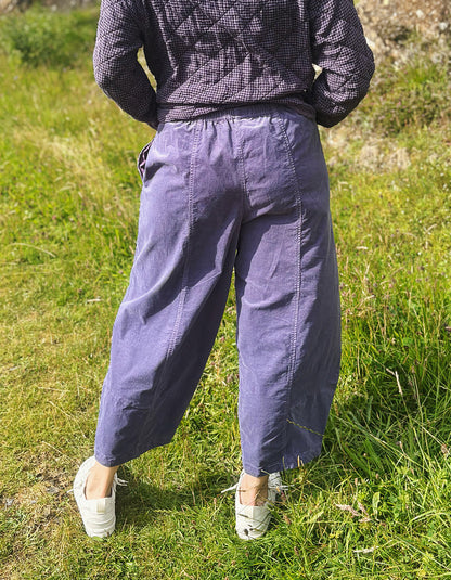 A person standing outdoors on grass, shown from the back wearing loose wide-leg pants in a muted dusty purple/lavender color, a dark patterned long-sleeve top, and white slip-on shoes.