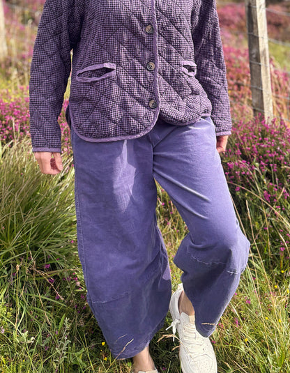 A person standing outdoors in a grassy field with pink flowers, wearing a quilted purple jacket and loose purple pants with white sneakers.