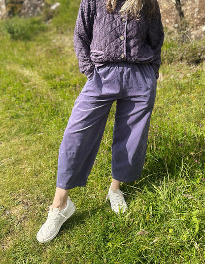 A person standing outdoors on grass wearing loose purple cropped pants, a matching quilted top, and white sneakers.