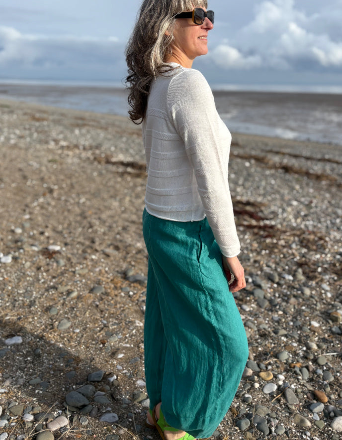 Woman standing on a pebbly beach wearing a white top and teal pants.