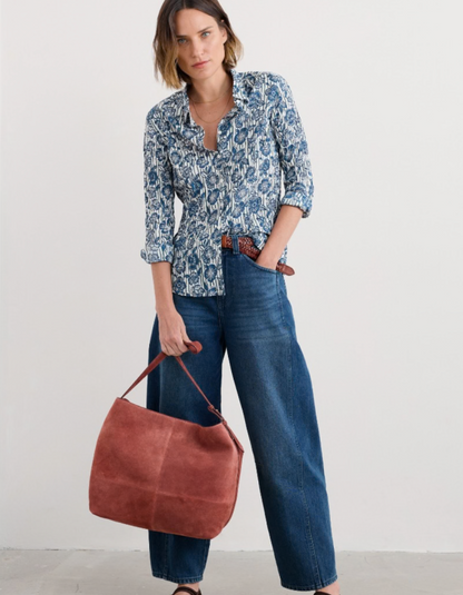 Woman wearing a blue patterned shirt and jeans, holding a brown leather bag on a white background