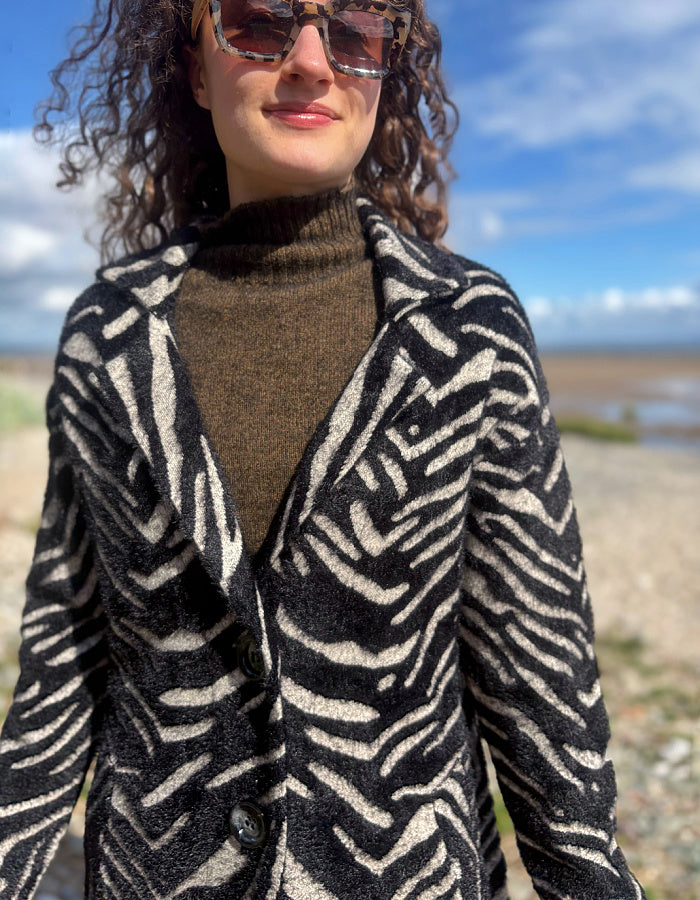 A woman outdoors wearing a black and beige zebra-print cardigan over a brown turtleneck sweater, with a beach and blue sky in the background.