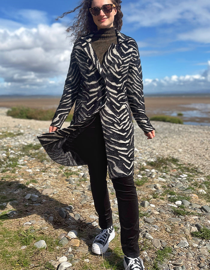 A woman wearing a black and white zebra-patterned open-front coat over a dark turtleneck, black pants, and black-and-white sneakers, standing outdoors on a rocky shoreline under a blue sky with clouds.