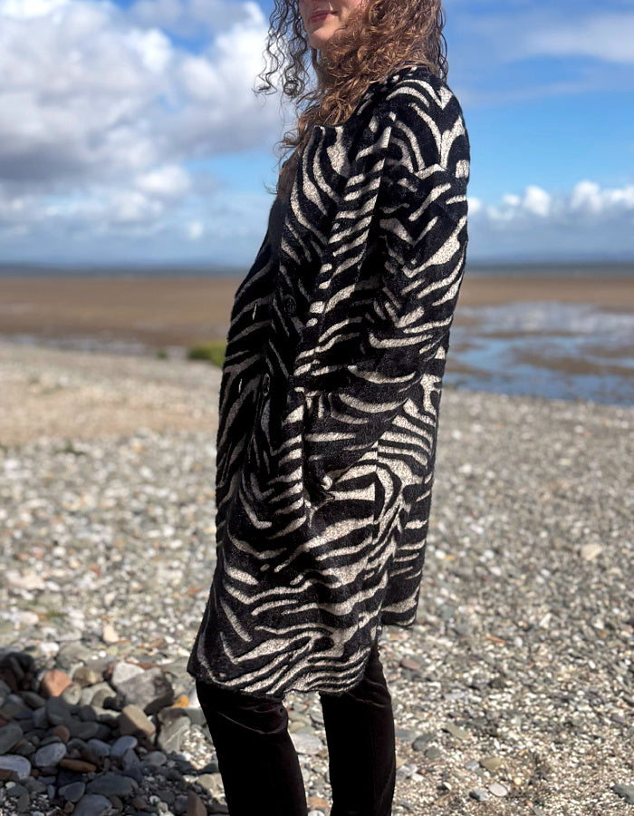A person standing on a pebble beach wearing a long black and off-white zebra-patterned cardigan or tunic, with dark leggings, against a background of water, sky, and distant land.