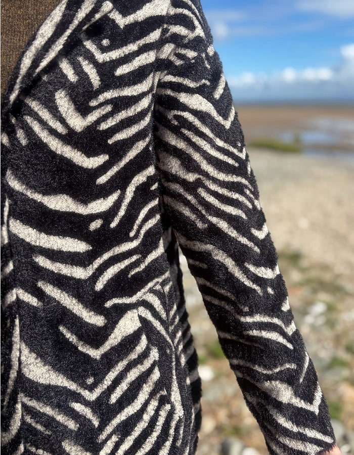 A close-up of a person wearing a black and beige zebra-patterned fleece jacket, with a blurred beach and blue sky background.
