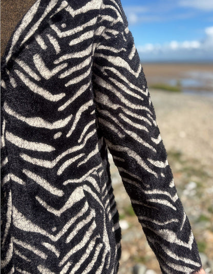 A close-up of a person wearing a black and beige zebra-patterned fleece jacket, with a blurred beach and blue sky background.
