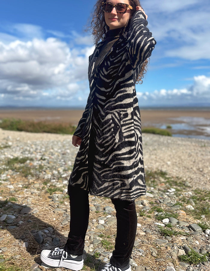 A woman wearing a black and beige zebra-print coat, black pants, and white sneakers stands on a rocky shoreline under a blue sky with clouds.