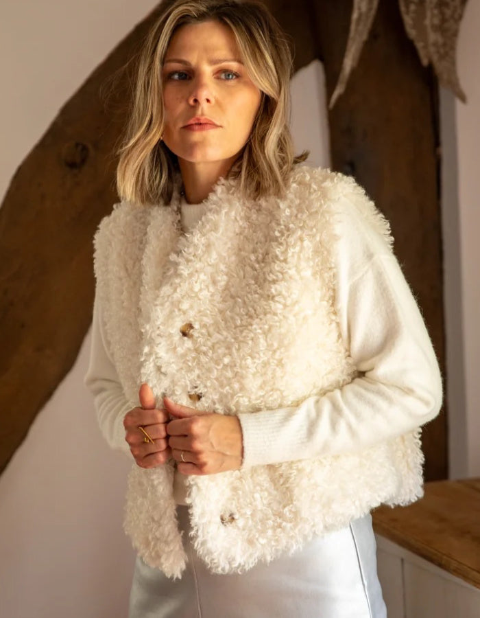 A woman wearing a cream-colored curly sheepskin vest over a matching cream knit sweater and white trousers, standing in a rustic room with a wooden beam background.