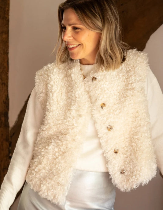 A woman wearing a cream-colored, curly-textured faux fur vest over a white sweater and white pants, standing in a rustic indoor setting.