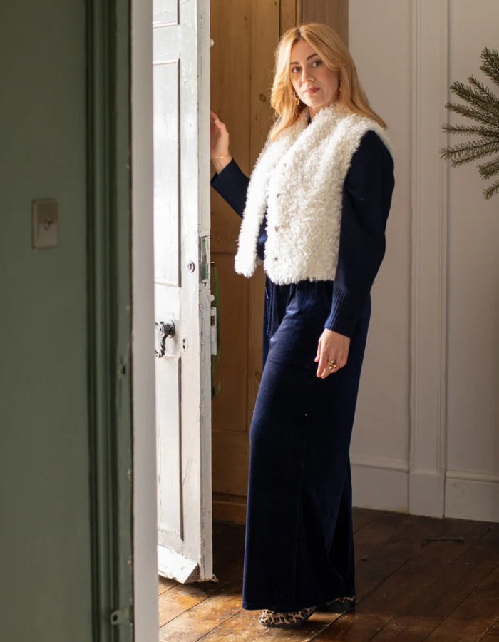 A woman stands in a doorway wearing a navy blue long-sleeve maxi dress with a white faux-fur vest layered over it. The photo is taken in a home interior with wooden floors, a white door, and a green wall.