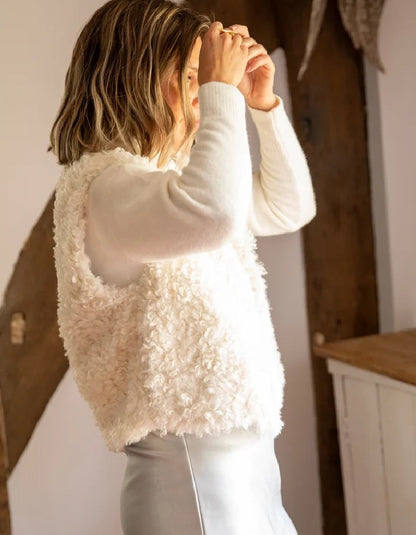 A woman wearing a cream-colored, fluffy textured vest over a white long-sleeve top and white pants, standing in a rustic indoor setting with wooden beams.