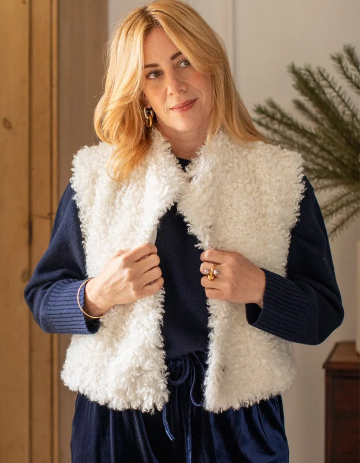 A woman wearing a white curly faux-fur vest over a navy blue ribbed sweater and matching navy skirt, standing in a warmly lit indoor setting.