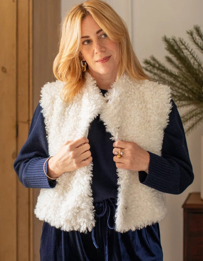 A woman wearing a white curly faux-fur vest over a navy blue ribbed sweater and matching navy skirt, standing in a warmly lit indoor setting.