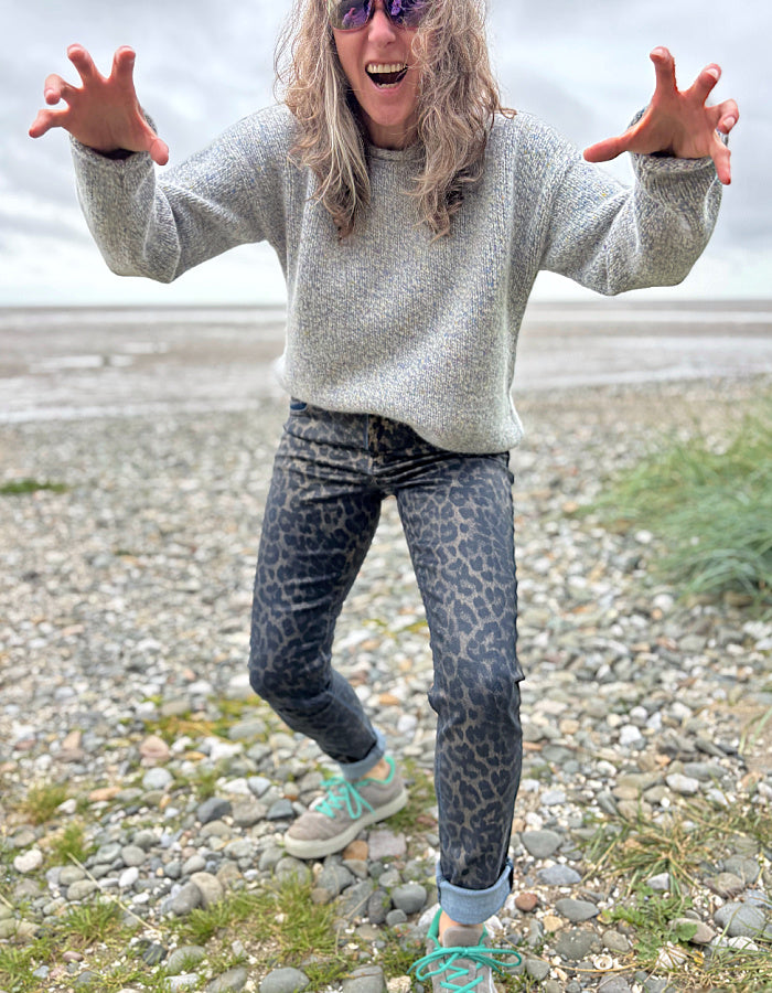 Person wearing a gray sweater and leopard print pants on a pebbly beach.