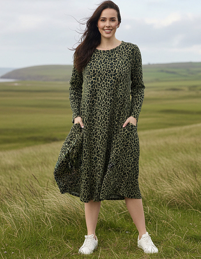 A woman wearing a long-sleeve olive green midi dress with a black leopard print pattern and white sneakers, standing in a grassy field with rolling hills under a cloudy sky.